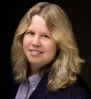 Noreen Grice Headshot - woman with blonde shoulder-length hair wearing a black blazer over a purple button up shirt