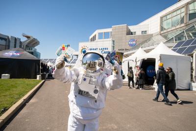 Someone stands wearing a complete space suit, white with shiny silver helmet, holding up the planet postcards and standing in front of Great Lakes Science Museum and NASA's Glenn Research Center