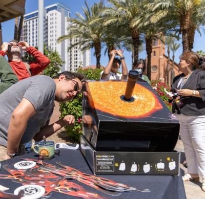 A sunny day for safe solar eclipse viewing, as palm trees sway in the back as a small crowd wearing eclipse glasses looks up in the background while a solar viewer site on a table with one person leaning in to look.