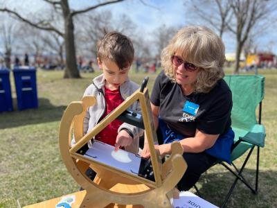 A volunteer uses a Sunspotter to help a young learner see and trace the shadow of the eclipse on paper using their finger.