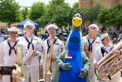 Five members of the Fort Worth Navy Band Southeast pose in white uniforms and eclipse glasses. They are holding their brass instruments while standing with uniform with Fort Worth Museum of Science and History mascot, Dynamo the Dinosaur.
