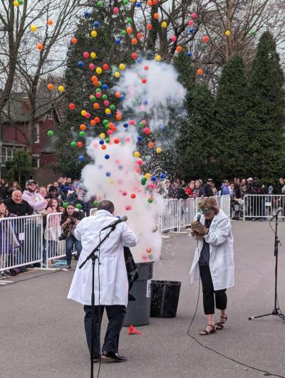 Science demonstration, two individuals in white lab coats stand near a large trash as a crowd watches white smoke and numerous multicolor balls fly out of the top.