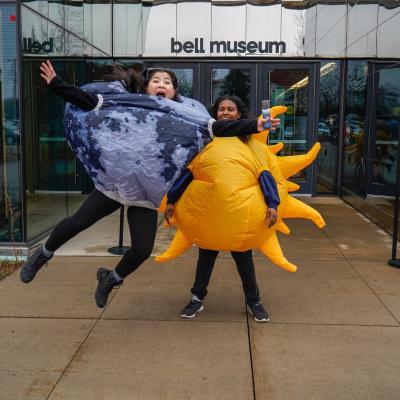 Two staff stand at The Bell Museum entrance and reenact the eclipse, one dressed as the Moon jumps in front to of the other dressed as the Sun to block it out.