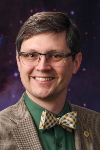 Smiling broadly enough to wrinkle the corners of his eyes, a light-skinned man with half-rim glasses and parted brown hair stands in front of a cosmic background. He wears a dark green shirt, a contrasting yellow and gray bowtie, a brown houndstooth jacket, and a lapel pin in the shape of the JWST primary mirror.