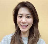 An individual from the shoulders up is smiling, their brown hair is swept over both shoulders and they are wearing a gray sweater.