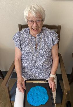 An elderly woman sits smiling with a tray on her lap. On the tray is a round lump of blue clay that has craters mimicking the surface of the Moon.