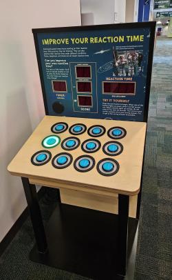 Standing exhibition component comprised of a large tombstone graphic with an attached table that is covered with 12 large blue buttons. The only visible text is "Improve Your Reaction Time"
