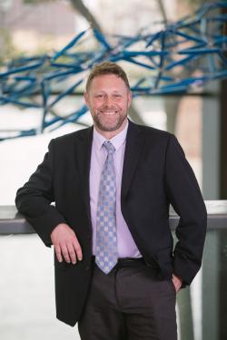 Man wearing a black suit with purple shirt and tie stands in front of a structure that is too blurry to see