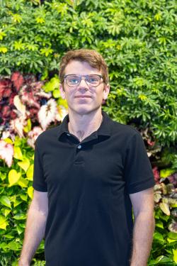 A man stands in front of a variety of colorful foliage. He is smiling while wearing glasses and a black shirt.