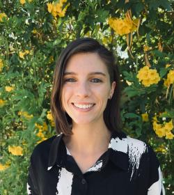 Closeup on an individual with dark shoulder-length hair, smiling and wearing a black and white top while posing in front of a large blooming yellow and green plant.