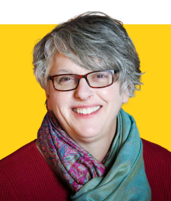 An individual from the shoulders up smiles, wearing a red shirt, green and purple scarf, and glasses. They have short grayish hair and appear on a yellow background.