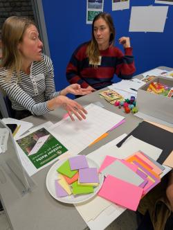 Two educators sit at a table with a variety of supplies such as sticky notes, hand outs, small toys and more.
