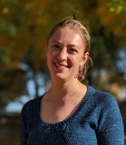 An individual poses smiling outside on a sunny day. They are wearing a blue sweater and have their hair up. Behind is a blurred background of greenery.