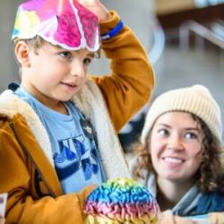 An adult kneels by a child at a table, on the table is a rainbow colored model of a human brain. The child is wearing a paper version of this rainbow brain on their head.