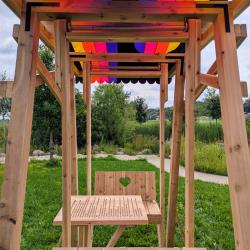 A wooden swing-like structure with a rainbow fabric roof, there is a small table with two seats right across from each other.
