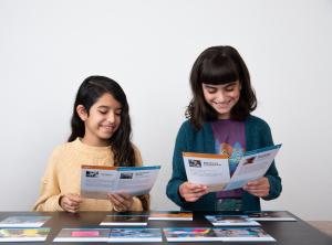 Two children are seated at a table spread with large cards that depict writing and various radio technologies. Both children are holding a card in each hand and smiling as they read the cards.