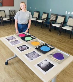 Educator stands by a table with multiple trays of brightly colored clay that has been flattened. By each tray is an image of the Moon's surface.