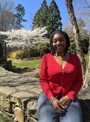 Outdoors on a sunny day, a woman sits on a stone wall with many different trees in the background. She is smiling, wearing a red shirt and blue jeans.
