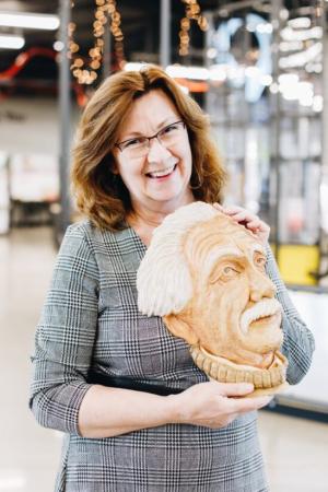 A women smiling and wearing glasses, she looks like she is in a museum and is holding a bust of Einstein.