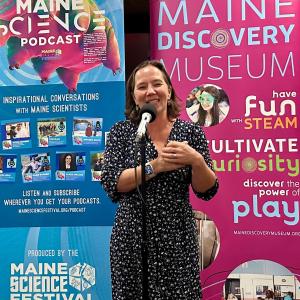 An individual wearing a black dress with white flowers stands at a mic smiling. It appears they are addressing a crowd we cannot see; behind is a wall with Maine Discovery Museum logos and branding.