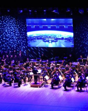 A full orchestra on stage, purple hues light their area while in the back a screen shows images of the Earth as seen from space.
