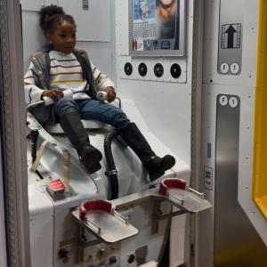 A child sits on a replica toilet for astronauts from the International Space Station. In the small white space, the child is strapped onto the toilet, with legs dangling because they are too short to reach the foot holds.