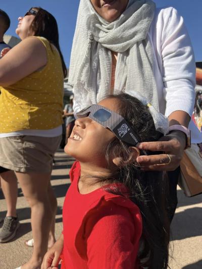 Child is viewing the eclipse while being aided by her Mother who is holding her eclipse glasses across her eyes.