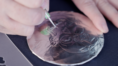 Close up of tin foil medallion as a pair of hands use a stylus to etch a sugar skull into the surface.
