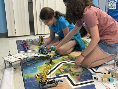 Two young individuals kneel on the floor with a few robotics items on a map with bold lines for the robots to follow.