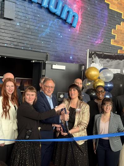 A small group stand smiling outside the door of a planetarium, where two of the individuals are holding large scissors and are about to cut a ribbon opening the space.