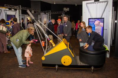 A youth kneels on a large round ottoman-like seat while watching a screen in front of him. This screen is part of a triangular exhibit with a long tube, and a much younger child and adult stand on the side with the tube loading it with small parachutes. 
