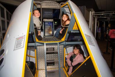Inside a space capsule exhibit, children sit inside on an upper deck while an adult sits below.