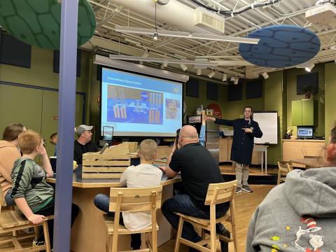 People sitting around multiple tables while watching a facilitator in a black lab coat present slides.