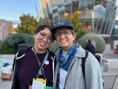 Two smiling people wearing conference badges and lanyards posing outdoors in front of a modern glass building.