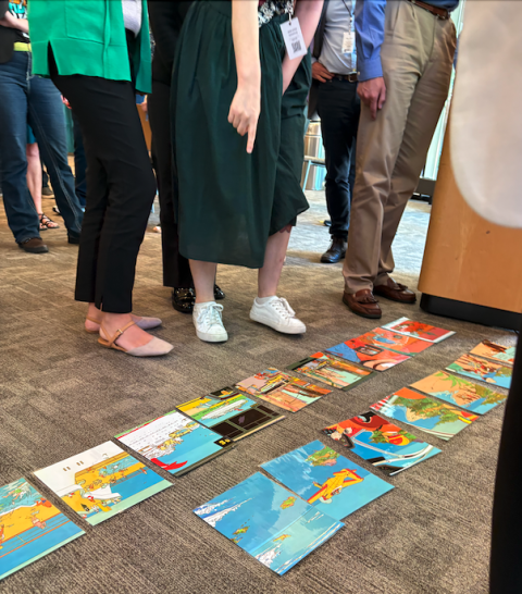 Multiple colorful images are spread across the floor in rows as a group of individuals seen only from the waist down stand and point at the images.