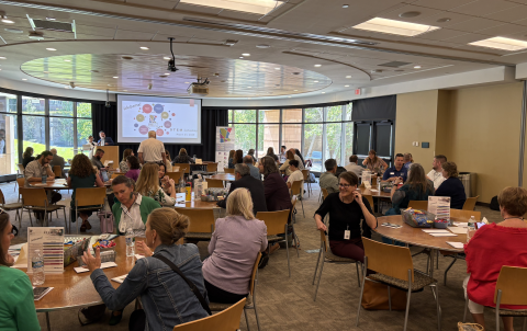 A large room set up for a presentation to begin, as many individuals sit at table rounds looking at a projection screen.