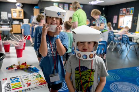 Two children pose wearing paper astronaut helmets while standing next to an activity station with the supplies to make the helmets.