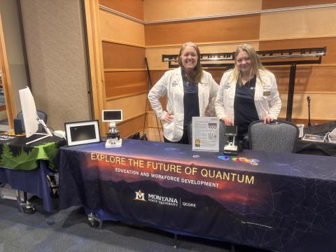 Two individuals stand by a table with a dark tablecloth that says Exploring the Future of Quantum. There is a sign and some small unidentifiable objects on the table.
