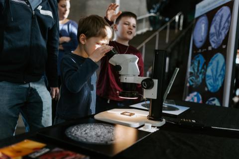 A child stands next to a table looking through some type of microscope. There are adults and other children standing around him who are also looking at images displayed around the table that look like enlarged microscope picture. 