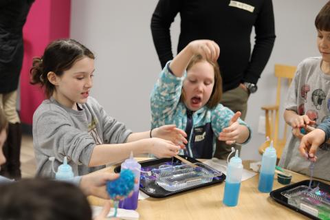 Two young people sit at a table, others are in the background. The two are both holding and manipulation a slime demo being done at the table.