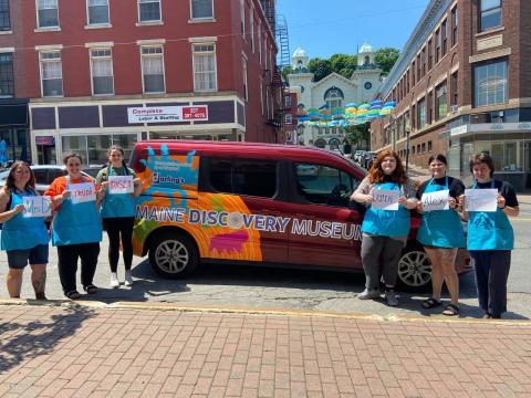 A group of six people all wearing the same light blue t-shirt stand next to a van. The van reads "Maine Discovery Museum" and each person is holding a white piece of paper with a name on it.