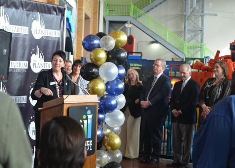 A speaker stands at a podium with a group surrounding them, standing amid a large display of balloons and exhibit components from Sun, Earth, Universe.