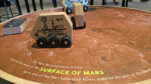 Closeup on a small round, wooden table with a tabletop graphic of Mars covering it and small wooden toy rovers on top.