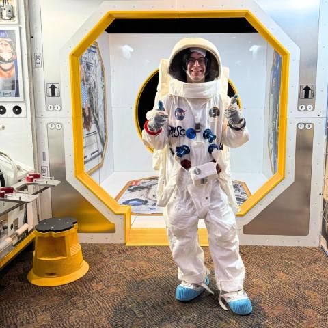 An individual wearing an astronaut costume gives two thumbs-up while standing inside an exhibit made to look like the International Space Station.