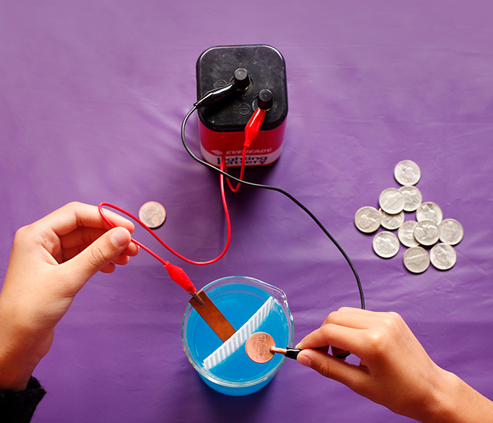 Hands holding wires connected to a battery over container of liquid in Exploring Fabrication - Electroplating activity.
