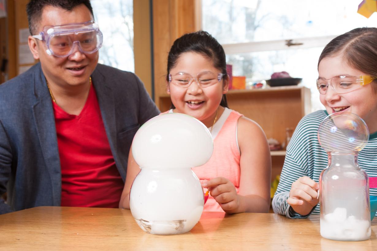 two learners and a facilitator watch a soapy solution with dry ice make a giant bubble filled with smoke