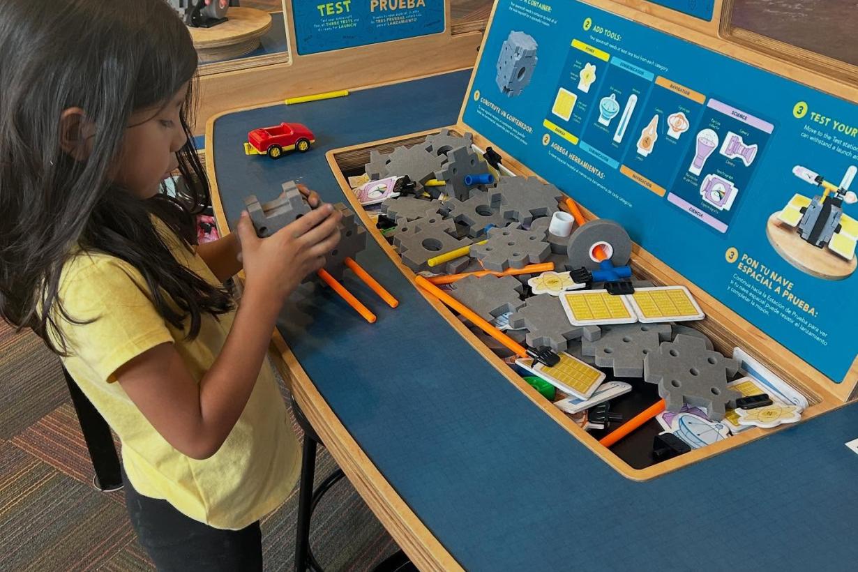 Children stand at opposite sides of the Design Build Test exhibit table, putting together the assorted pieces and testing their creations.