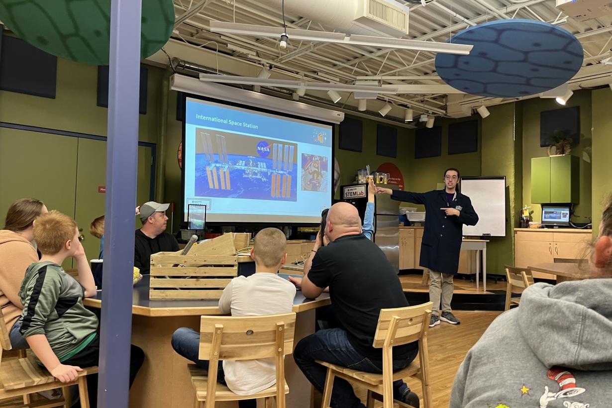 People sitting around multiple tables while watching a facilitator in a black lab coat present slides.