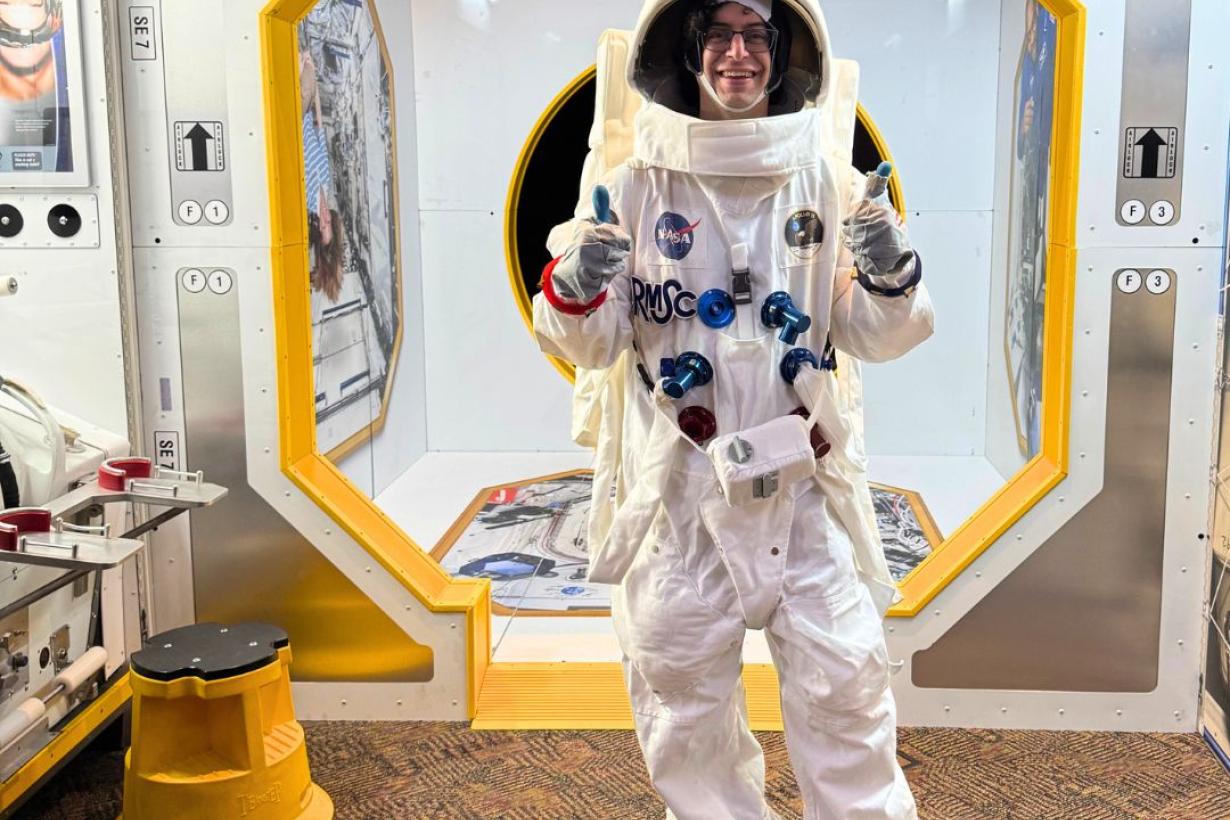 An individual wearing an astronaut costume gives two thumbs-up while standing inside an exhibit made to look like the International Space Station.