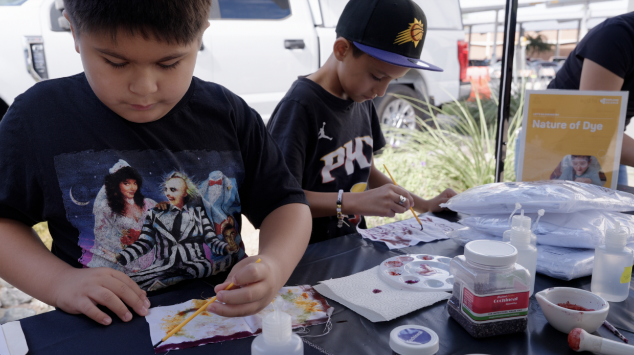 Two young kids stand at a table with paints, brushes, and what looks like thin fabric to paint on. They have heads down, focused on the painting activity 